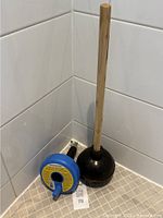 Toilet plunger and PlumbShop drum auger positioned side by side in a tiled corner, showing the plunger's wooden handle and black rubber suction cup.