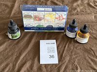 Photo of original sealed box with 8 Winsor & Newton Drawing Inks bottles, arranged behind loose bottles of Sennelier ink on a brown fabric background