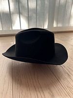 Black wool hat displayed on wooden floor in front of window, showing slight shape distortion but intact brim and crown structure.