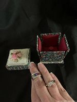Two rings worn on fingers and opened decorative floral jewelry box showing red velvet interior and ring holder insert.