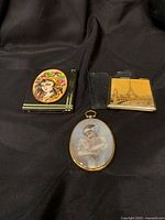 Three vintage compact mirrors displayed on black background showing Eiffel Tower scene, lady with flowers, and oval mirror with girl holding flowers