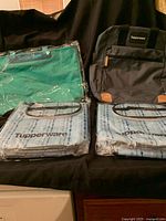 Four Tupperware bags displayed on a black surface: two light blue insulated bottle totes in plastic packaging, one green shopping tote, and one gray backpack.