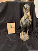 Overall view of both items in the lot: framed parakeet lapidary art and carved wood heron sculpture on black backdrop.