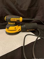 Side view of yellow and black DeWalt corded random orbital sander with dust collection bag attached, laid on white and black background.