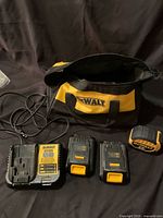 Dewalt carrying bag, charger, two batteries, and tape measure laid out on dark surface