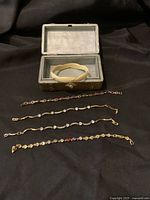 Lot of six gold-tone bracelets and one bangle displayed on a black cloth with vintage jewelry box open showing gray velvet interior.