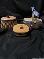 Three Longaberger handwoven wood baskets with fitted wooden lids and fabric liners on black fabric background, arranged side by side.