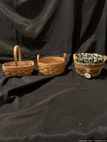 Photo showing three small woven baskets arranged side by side on a black background: a basket with a single wooden handle, a basket with two leather handles, and a basket with decorative fabric liner and tag.