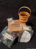 Miniature Longaberger basket with fabric liner, floral tie-on, plastic protector, and original box on black background, items still in plastic packaging.