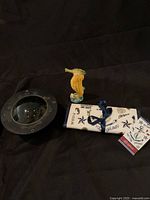 Photo showing the Brighton travel jewelry pouch rolled and tied with ribbon, vintage ships’ compass with black metal housing and glass dome, and a ceramic seahorse figurine with detailed glaze.