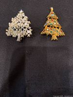 Photo of front view of two Christmas tree brooches, one silver-tone with clear rhinestones, one gold-tone with multicolored rhinestones.