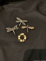 Four brooches displayed on black fabric background, showing three dragonfly pins and one circular floral pin.