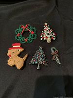 Five Christmas-themed brooches on black background including a green tatted wreath with red bows, two Christmas tree brooches, a candy cane brooch, and a wooden dog brooch.