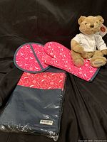 Photo showing red Tupperware oven mitt and matching pot holder with blue trim, featuring a pattern of kitchen icons and logos. The items appear to be made from cotton fabric. The pot holder is still in original plastic packaging.