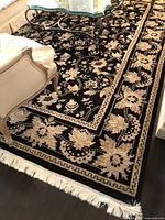 Corner of rug showing black ground, ivory floral border and fringe on one short edge under living room chair.