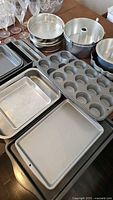 Photo showing rectangular cookie sheets, metal muffin tins, and cake pans arranged on tabletop with visible wear.