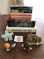 Stack of six books topped with a Premier Dominoes box and surrounded by four small decorative figurines on a table.