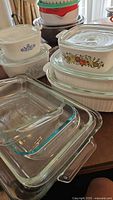Stacked assortment of clear glass Pyrex casserole dishes and CorningWare ceramic casseroles with lids, showing vintage fruit and blue floral patterns.