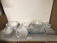 Full set showing three Corningware casserole dishes with blue cornflower pattern and their glass lids, one white Corningware rectangular baking dish with green floral pattern, two clear glass mixing bowls, and two clear glass baking trays.