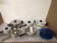 Full view of all cookware including the blue Dansk Kobenstyle pot, three white floral enamel pots, and three stainless steel pans.