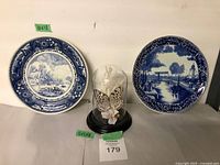 Two round Delft blue and white plates flanking a glass dome with butterfly, shown on a flat surface