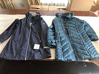 Two coats laid flat on a table, showing the front sides: a dark blue patterned hooded spring coat and a teal quilted hooded winter coat.