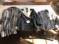 Three ladies jackets laid out flat showing styles and colors: gray plaid cardigan, black double-breasted with white lapel, and black and white patterned jacket.