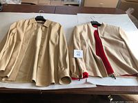 Two beige ladies jackets on hangers laid flat on a table; one is a leather button-up shirt and the other a zip jacket with red interior lining.