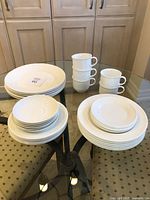 Full view of complete dish sets including stacked dinner plates, side plates, shallow bowls, and cups with handles arranged on glass table.