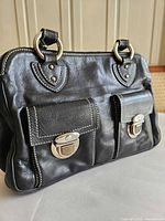 Angle view of the black leather purse showing two front flap pockets with silver-tone turn-lock closures and rounded handles attached with silver-tone rings.