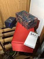 Photo of three decorative boxes stacked; includes blue and floral patterned boxes and large red satin-covered box.