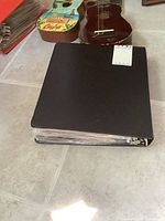 Photo of a large black binder filled with plastic-protected comics placed on the floor next to two ukuleles.