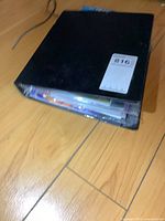 Closed black binder lying on wooden floor, showing some wear along edges and filled with comic books.