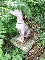 Large concrete dog statue in seated position, mounted on a heavy rectangular concrete base surrounded by greenery. Shows signs of wear and paint loss.