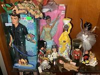 Photo showing various dolls and figures including a boxed male My Scene doll, dolls in cultural costumes, a ballerina doll, and other small dolls.