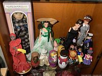 Full view of assorted dolls and figures on shelf, including boxed porcelain doll, fashion dolls, and wooden nesting dolls