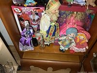 Dolls and figures arranged on wooden shelf including the boxed Dale Jr NASCAR Barbie and multiple fabric and traditional dolls.