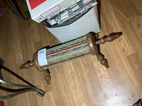 Overall angled view of the vintage wooden foot rest on hardwood floor with multicolored carpet wrapped around cylinder body and turned wooden ends and legs.