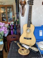 Photo showing three items: a full-size child’s acoustic guitar standing vertically, a small toy banjo lying flat on a table, and a small toy violin partly visible beside the banjo.