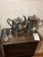 Two silver plated teapots of hexagonal shape on a wooden table alongside two Hoselton metal figures.