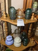Collection of nine different vintage glass and porcelain insulators displayed on two-tiered wood stand. Various colors and styles visible including green, clear, blue, white and ridged stoneware base insulator.