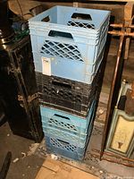 Stack of four plastic milk crates, three blue and one black, showing lattice design, handles, and size relative to surroundings.