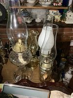 Four vintage glass oil lamps of varying sizes sitting on a wooden surface with shelves of ceramics and other items behind them. Lamps have glass chimneys, mostly clear, one frosted white.