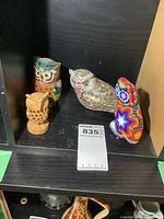 Four owls on a black shelf: wooden carved owl, multicolor ceramic/resin owl, realistic feathered resin owl, multicolor beaded owl