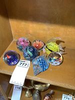 Seven glass paperweights displayed on a wooden shelf, showing a variety of shapes and internal designs including fish and floral motifs.