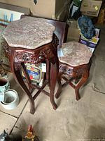 Pair of wooden plant stands with rose carvings and scalloped pink marble tops, one taller and one shorter, photographed together on a cardboard floor in storage area.
