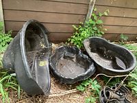 Three black plastic garden ponds placed outdoors against wooden fence, showing varying irregular shapes and wear.