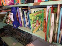 Photo showing multiple children's pop-up books on a green shelf with visible titles including Little Red Riding and Dinosaur Pop-Up.