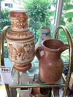 Three pottery pieces including decorative relief vase, rustic brown pitcher, and a white and blue vase/pitcher on a glass shelf with window behind.