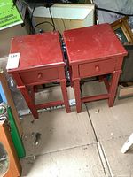 Pair of small red wooden side tables with single drawers, placed side by side showing tops and legs with wear and minor scuffs.